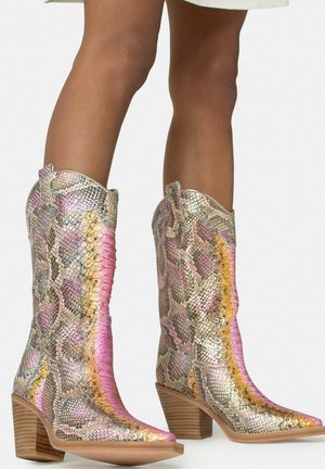 Piernas con botas de vaquero hasta la mitad de la pantorrilla, con punta afilada, tacón apilado de madera y patrón de piel de serpiente en rosa, amarillo y beige.