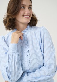 Blouse bleu clair avec un col à volants, motif zigzag texturé et manches plissées élastiques. Boutonnage sur le devant.