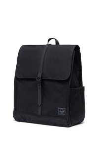 Crni platneni ruksak s preklopom, podesivim kožnim remenom, bočnom džepom i teksturiranom površinom. Ima oznaku loga na prednjoj strani.