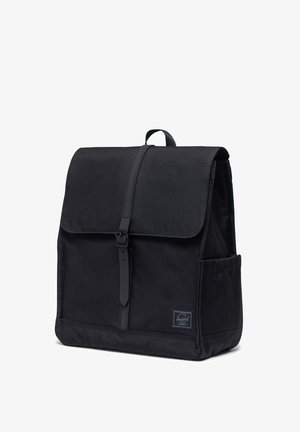 Mochila de tela negra con cierre de solapa, correa de cuero ajustable, bolsillo lateral y un acabado texturizado. Presenta un parche con el logotipo en la parte delantera.