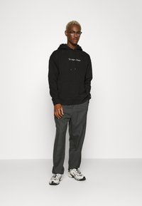 Felpa nera con il logo testuale "Tommy Jeans", pantaloni grigi e sneakers bianche. Il materiale sembra morbido; la felpa ha una tasca a marsupio frontale.