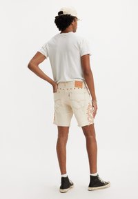 Shorts de mezclilla beige con bordes deshilachados, con bordado floral naranja en el lado izquierdo, combinados con una camiseta gris y zapatillas negras.