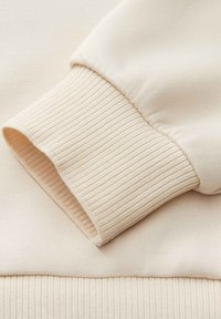 Cream-colored sweatshirt detail featuring a ribbed cuff and smooth texture, highlighting the fabric's soft finish and seamless attachment.