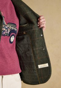 Persoon die een roze trui draagt met een bloemmotief auto-ontwerp houdt een groene geruite tweed jas open en toont de patroonvoering en het label.