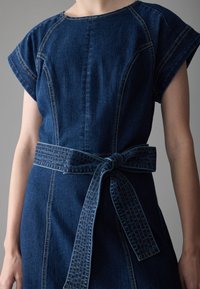 Vestido de mezclilla de longitud media en azul oscuro, con mangas cortas, una cintura ceñida con un cinturón atado, detalles de costura visibles y una textura suave.