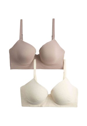Donna con capelli ricci solleva un maglione beige per rivelare un reggiseno bianco senza cuciture e senza spalline e biancheria intima coordinata su sfondo bianco.