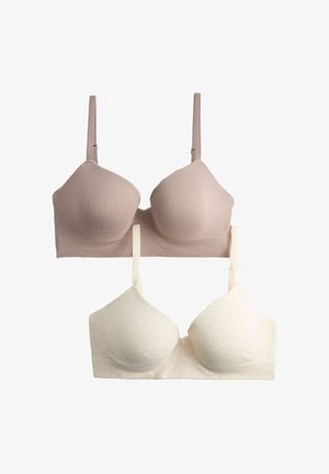 Femme aux cheveux bouclés soulève un pull beige pour révéler un soutien-gorge blanc sans coutures ni bretelles et un sous-vêtement assorti sur fond blanc.