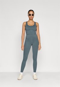 Tielový športový outfit z texturovanej tkaniny, ktorý obsahuje tielko a legíny s vysokým pásom. Doplnkovaný bielymi športovými topánkami a slnečnými okuliarmi.