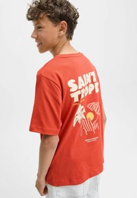 Camiseta de algodón roja con estampado gráfico "SAINT TROPEZ" en la parte posterior, que incluye ilustraciones de palmeras, un sol y una silla de playa. Manga corta.