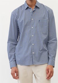 Hombre con una camisa de cuadro azul y blanca de manga larga y pantalones beiges, de pie contra un fondo blanco.
