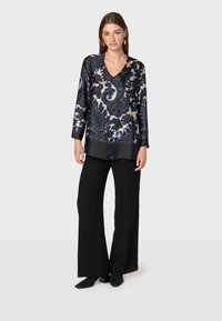 Maglia a maniche lunghe con scollo a V, caratterizzata da un motivo paisley crema e blu, abbinata a pantaloni larghi neri e calzature nere.