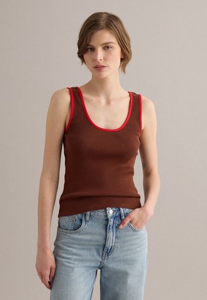 Jeune femme aux cheveux courts et bruns portant un haut marron côtelé sans manches avec une bordure rouge et un jean bleu clair, debout devant un fond uni.