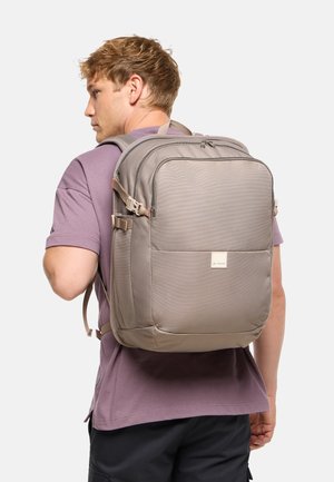 Homme portant un sac à dos rectangulaire marron clair avec plusieurs compartiments et des boucles latérales, vêtu d'un t-shirt violet, sur un fond uni.