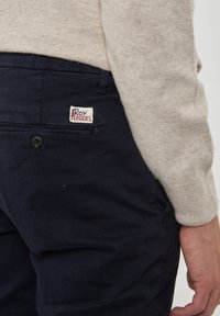 Pantaloni di cotone blu navy con tasca con bottone, con etichetta che riporta "Roy Rogers". Il tessuto ha una consistenza liscia e un design a taglio dritto.