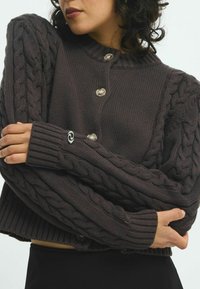 Virkat kavaj i mörkgrått med kabelstickade ärmar, rund hals och stor knapparstängning. Strukturerad, kortad passform med ribbade handskar.