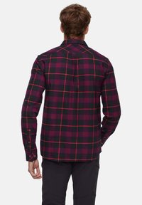Uomo visto da dietro che indossa una camicia a maniche lunghe a quadri neri e viola scuro con linee rosse e pantaloni neri, contro uno sfondo neutro.