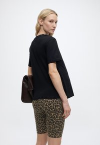 Γυναίκα με ξανθά μαλλιά φοράει μαύρο κοντομάνικο μπλουζάκι, σορτς ποδηλασίας με λεοπάρ μοτίβο και κρατά σκούρα καφέ δερμάτινη τσάντα σε μονόχρωμο φόντο.