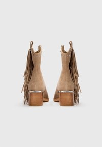Botas de tobillo de ante marrón claro con tacón de bloque de madera, que presentan detalles de flecos y tachuelas plateadas a lo largo del borde superior.