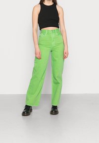 Jeans verdes de pierna ancha con cintura alta, hechos de algodón. Combinados con una camiseta sin mangas negra y unos zapatos negros con cordones.