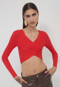 Frau mit langen dunklen Haaren, die ein rotes geripptes Wickel-Crop-Top, große goldene Ohrringe und dunkelbraune Lederhosen trägt, posiert mit den Händen in den Taschen.