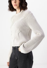 Kremast pulover iz pletene tkanine z rebrasto teksturo, okroglo ovratnico in spuščenimi ramenom, v kombinaciji s črnimi denim hlačami z stranskim žepom.