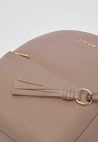Sac rond en similicuir beige avec une poche zippée, des éléments dorés et un détail de pompon décoratif. Présente un logo de marque embossé "LIUJO".