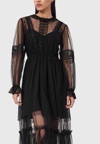 Μαύρο φόρεμα από διάφανο πουά ύφασμα με μακριά μανίκια, δαντελένιες λεπτομέρειες, βολάν στο λαιμό και στρώσεις στο τελείωμα. Διαθέτει εφαρμοστό μπούστο.