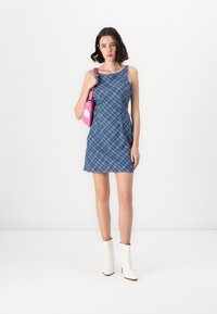 Modra karirana brezrokavnica iz denima, ki ima prilegajočo obliko. V kombinaciji z sijočo roza torbico in belimi gležnjači.