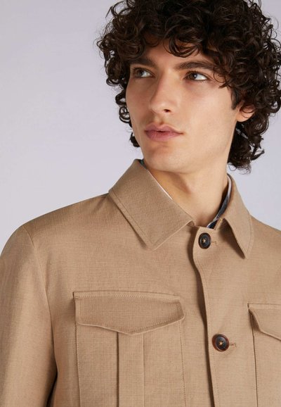 Beige Jacke aus strukturiertem Stoff, mit spitzem Kragen, zwei Brusttaschen und braunen Knöpfen.