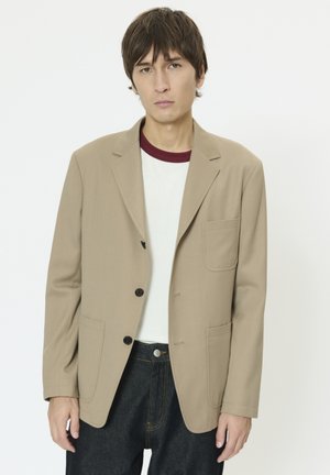 Ung mand med kort brunt hår iført en beige blazer over en hvid t-shirt med mørkerød krave og mørkeblå jeans, stående mod en hvid baggrund.