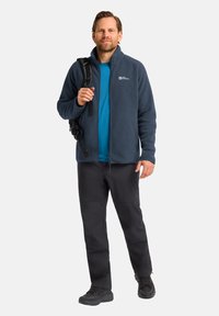 Giacca in pile blu sopra una camicia blu, pantaloni neri e scarpe da trekking nere. Persona che tiene uno zaino nero con una tracolla.