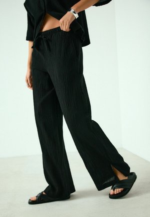 Woman wearing black textured wide-leg pants with side slits and black slip-on sandals, standing on a light floor against a white wall.