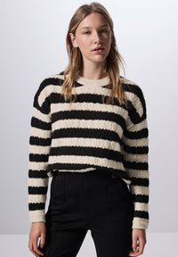Gestreifter Pullover in Schwarz und Creme mit strukturiertem Strickdesign. Verfügt über einen runden Halsausschnitt und lange Ärmel, kombiniert mit schwarzen Hosen.