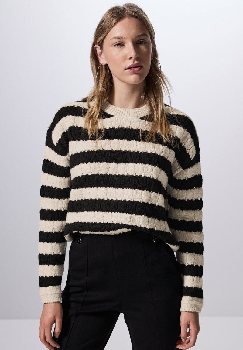 Gestreifter Pullover in Schwarz und Creme mit strukturiertem Strickdesign. Verfügt über einen runden Halsausschnitt und lange Ärmel, kombiniert mit schwarzen Hosen.