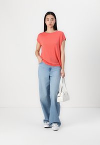 Koral kortermet t-skjorte kombinert med lyseblå vide jeans og hvite sneakers, og holder en liten hvit håndveske. Glatt stoff og komfortabel passform.