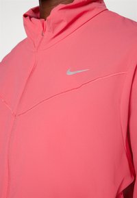 La veste à capuche rose Nike présente une fermeture éclair, une texture lisse et un logo argenté sur la poitrine. Conçue pour un confort léger.