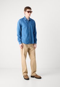 Camicia di jeans button-up blu, pantaloni khaki e scarpe marroni; il modello indossa occhiali da sole marroni. La texture appare liscia, con una vestibilità casual.
