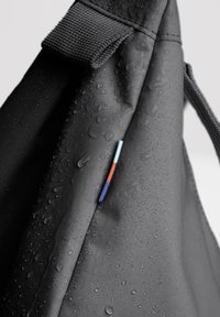 Zaino impermeabile nero con superficie testurizzata. Presenta un piccolo tag colorato e gocce d'acqua sul tessuto.