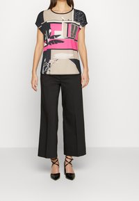 Top a maniche corte con un motivo geometrico in beige, rosa e nero, abbinato a pantaloni larghi neri e scarpe con tacco nere.