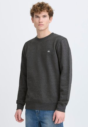 Sweatshirt gris avec un col rond, des poignets et un ourlet côtelés. Présente un petit logo blanc sur la poitrine gauche. Porté avec un jean bleu.
