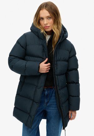Marine pufferjack met een capuchon, voorzien van geborduurde secties en een rits. Draagt over een zwarte trui en blauwe jeans.