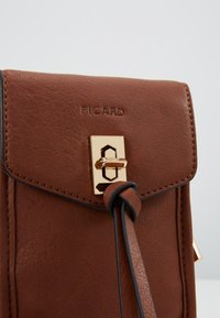 Borsa in pelle marrone con chiusura a scatto rettangolare dorata e nodo del cinturino bordato di nero sul flap anteriore, embossata con il nome del marchio "PICARD".