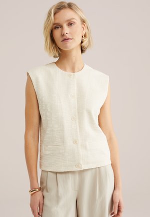 Donna bionda che indossa un gilet bianco senza maniche con bottoni e tessuto strutturato, pantaloni beige, accessoriati con braccialetto d'oro e orecchini a cerchio.