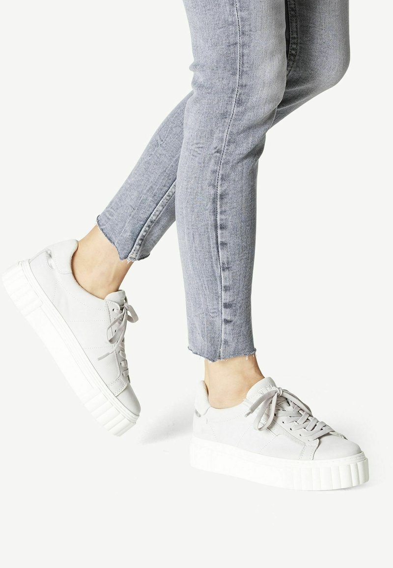 Vaaleanharmaat tennarit, joissa on teksturoitu yläosa, litteät nauhat ja paksu valkoinen platform-pohja, yhdistettynä kuluneisiin farkkuihin, jotka on leikattu nilkkaan.