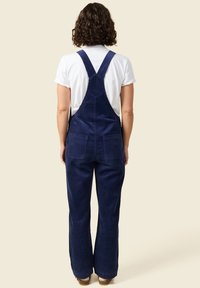 Marineblaue Corduroy-Latzhose mit weitem Bein, verstellbaren Schulterriemen, zwei Gesäßtaschen und einer glatten Textur, getragen über einem weißen T-Shirt.