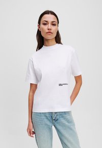 Balta medvilninė marškinėliai su apvaliu iškirpimu ir trumpomis rankovėmis, ant apatiniame kairėje pusėje esantis juodas tekstinis logotipas "KARL LAGERFELD JEANS".