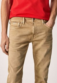 Jeans de algodón beige con corte ajustado, que cuentan con cinco bolsillos, acentos de metal y un acabado de lavado ligero. Combinados con una camisa roja.