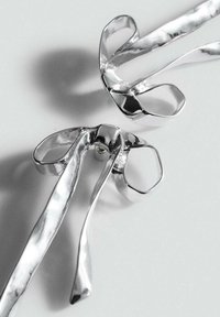 Clip a forma di nastro in metallo argento con una texture martellata e estremità ad anello, caratterizzati da una finitura liscia e riflessi delicati.