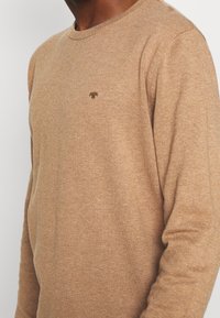 Mężczyzna w jasnobrązowym swetrze z dzianiny, z małym ciemnobrązowym haftowanym logo po lewej stronie klatki piersiowej.