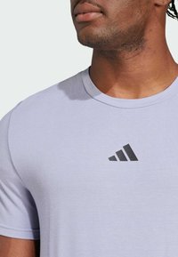 Camiseta de manga corta color lila claro, hecha de una tela suave, con un logo triangular negro de Adidas en el lado izquierdo del pecho.